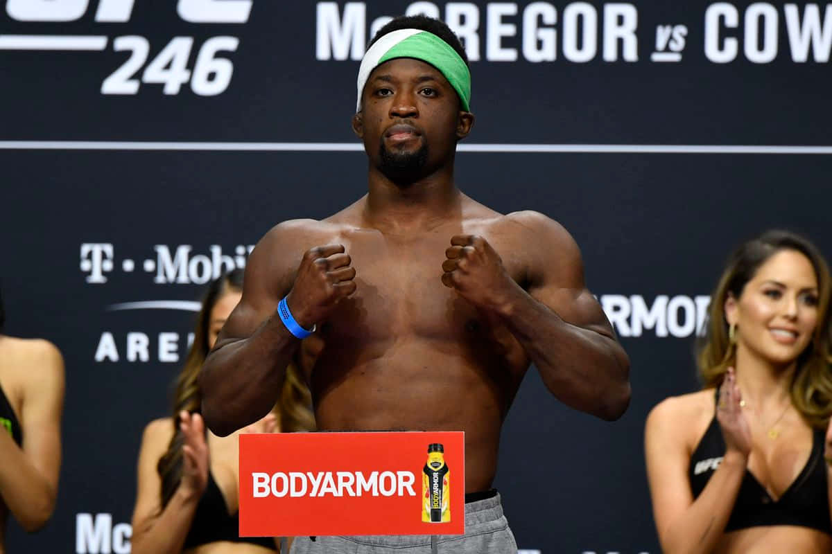 UFC 桌布 壁紙 — 一名肌肉發達、戴著綠色頭帶的拳手在UFC稱重活動中自信地擺出姿勢，背景有品牌標誌和歡呼的隨行人員。