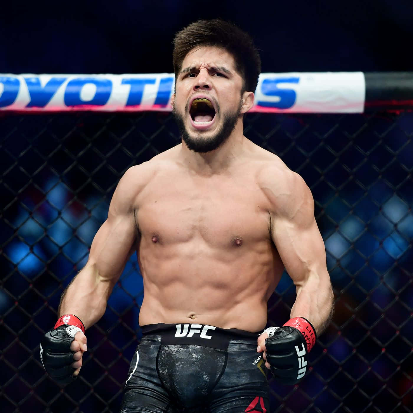 UFC 桌布 壁紙 — 一名肌肉發達的UFC拳手在八角籠內勝利咆哮，身後是模糊的人群和明亮的燈光。