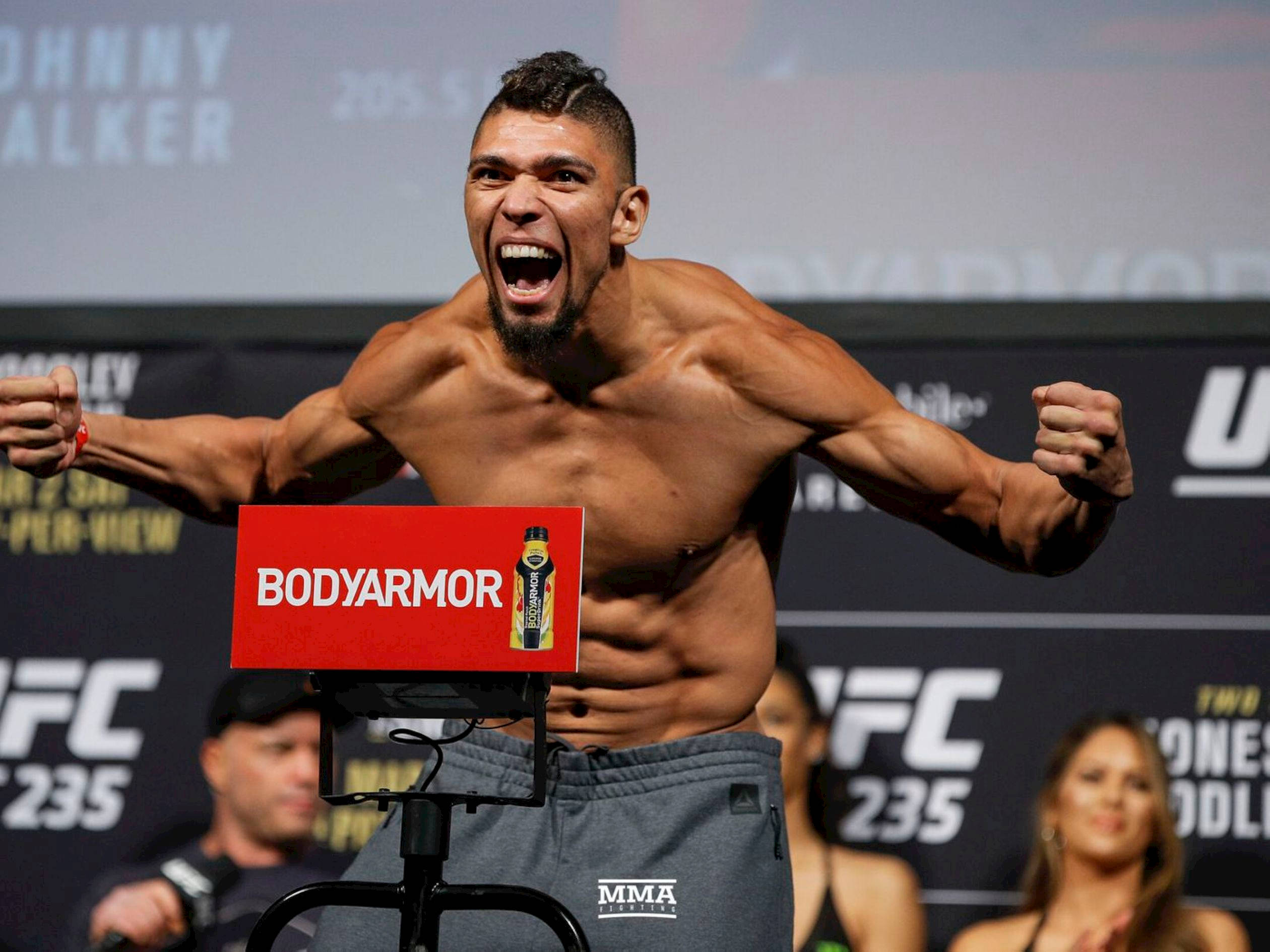 UFC 桌布 壁紙 — 一名肌肉發達的UFC拳手在稱重活動中，站在體重計上高舉拳頭，發出勝利的吼叫。