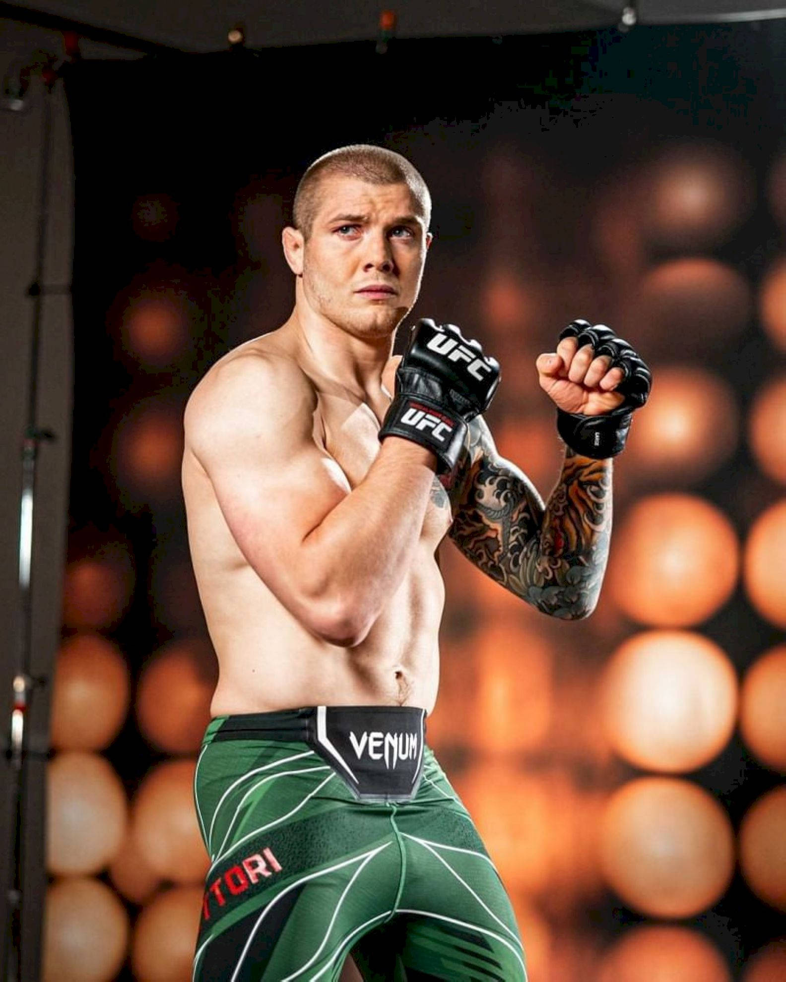 UFC 桌布 壁紙 — 一名肌肉發達的UFC拳手穿著綠色短褲和黑色手套，以戰鬥姿勢站立，手臂上可見刺青，背景是模糊的橙色燈光。