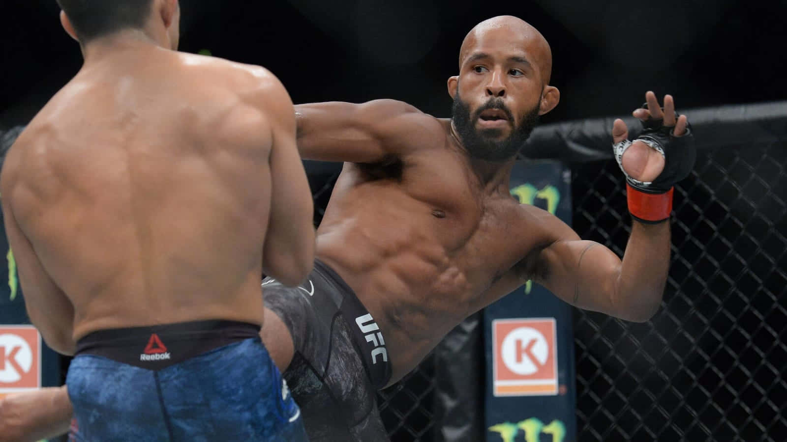 MMA 桌布 壁紙 — 一名肌肉發達的UFC拳手穿著黑色短褲，在燈光昏暗的八角籠內向對手揮拳。