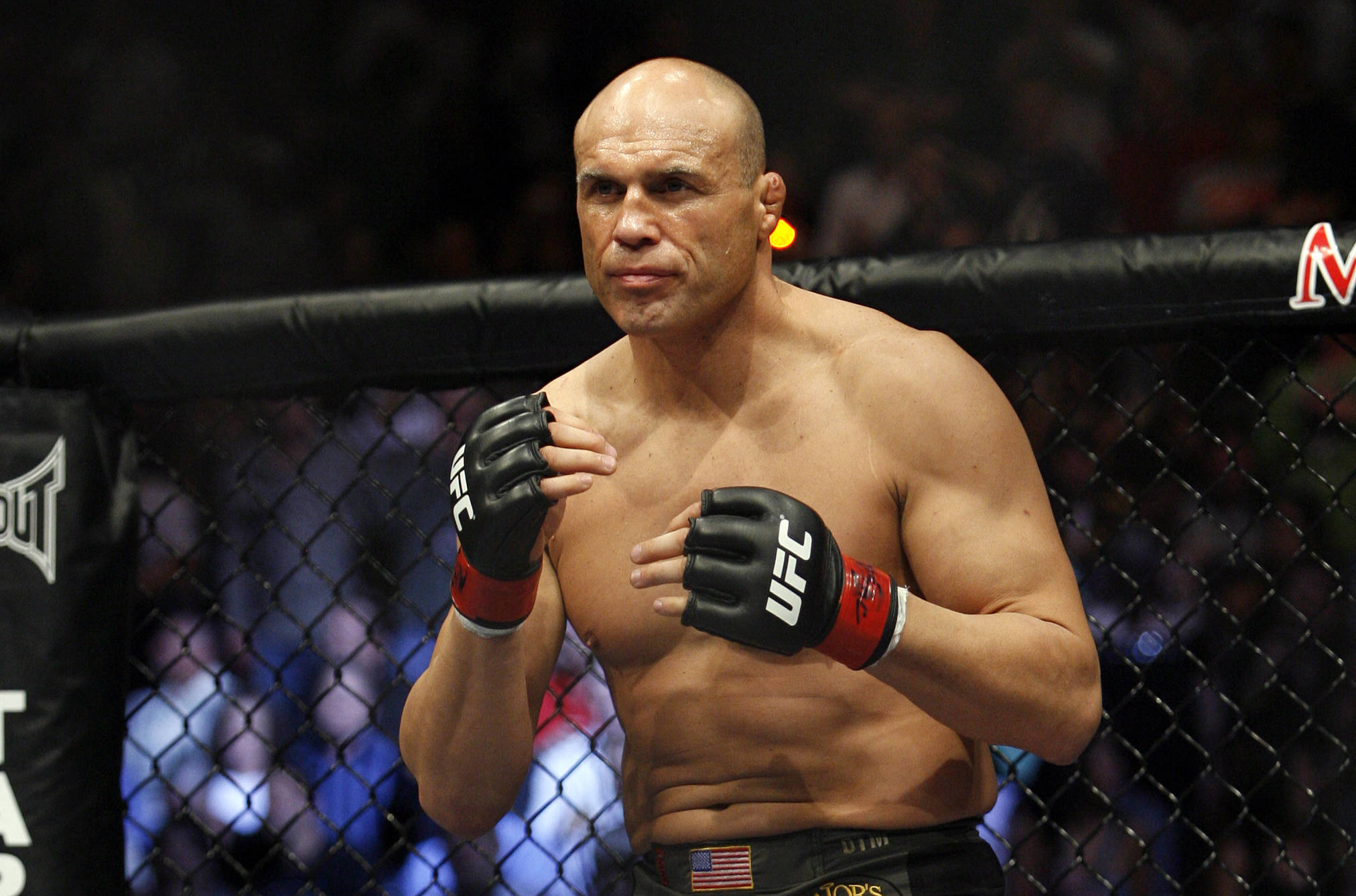 MMA 桌布 壁紙 — 一名肌肉發達的光頭拳擊手戴著UFC手套，在八角籠內警覺地站立，背景是模糊的人群。