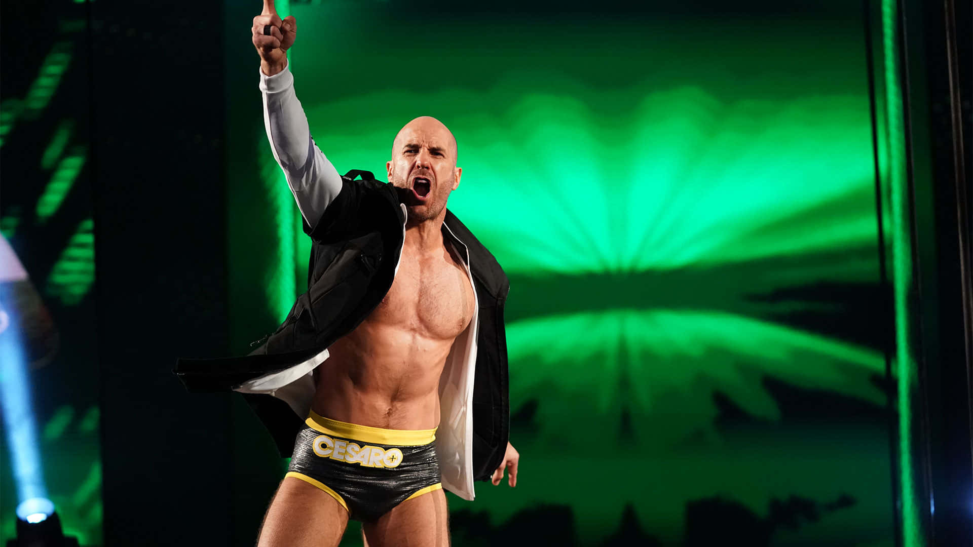 Swiss 桌布 壁紙 — 一名肌肉發達的光頭摔角手，穿著印有「CESARO」的黑黃色短褲，赤裸上身，外套敞開，在充滿活力的綠色舞台背景下勝利地舉起右拳。