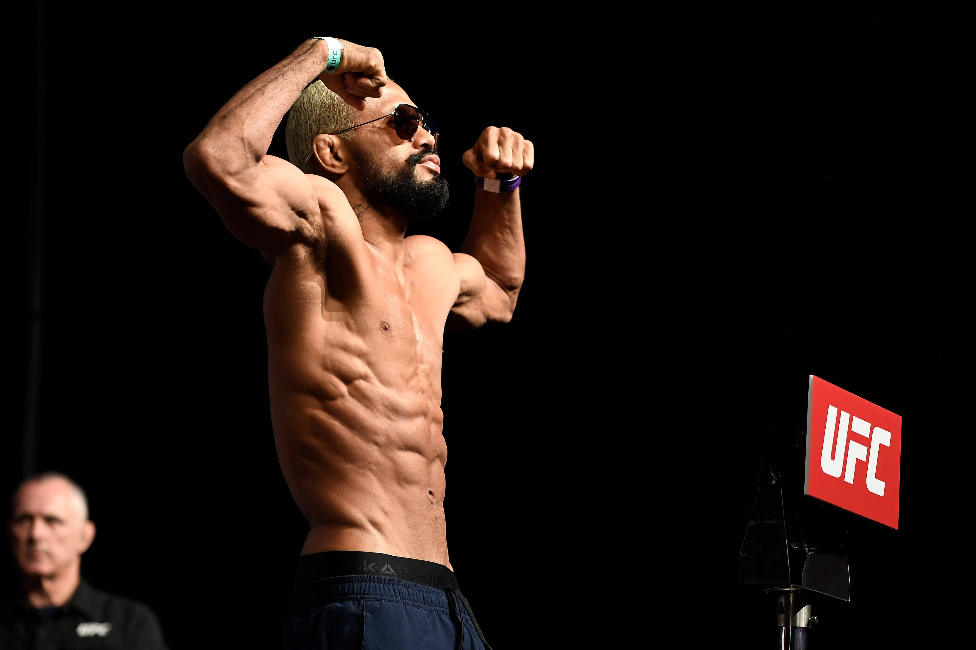 MMA 桌布 壁紙 — 一名肌肉發達的戰士在明亮燈光下繃緊腹肌，身後有UFC標誌發光。