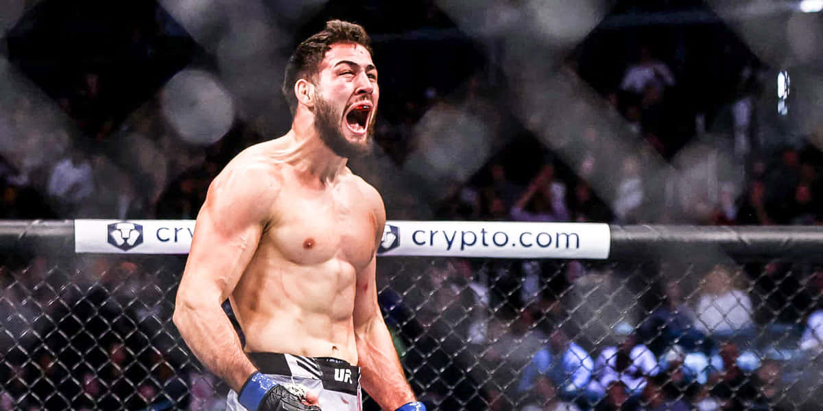 UFC 桌布 壁紙 — 一名肌肉發達的戰士在燈火通明的八角籠內咆哮勝利，圍欄後是模糊的人群。