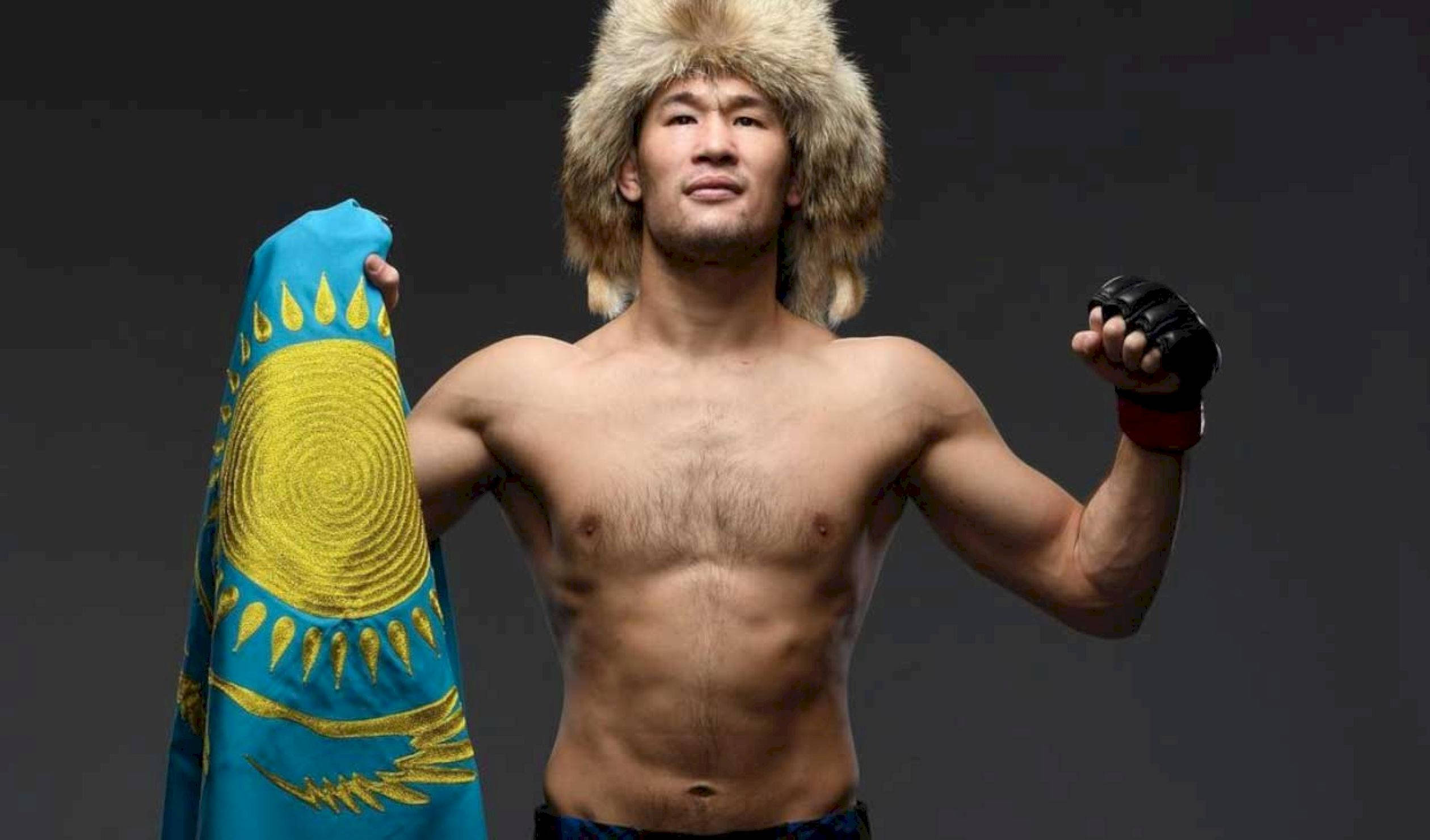 UFC 桌布 壁紙 — 一名肌肉發達的戰士戴著毛帽，在黑暗的攝影棚背景下自豪地舉起哈薩克國旗。