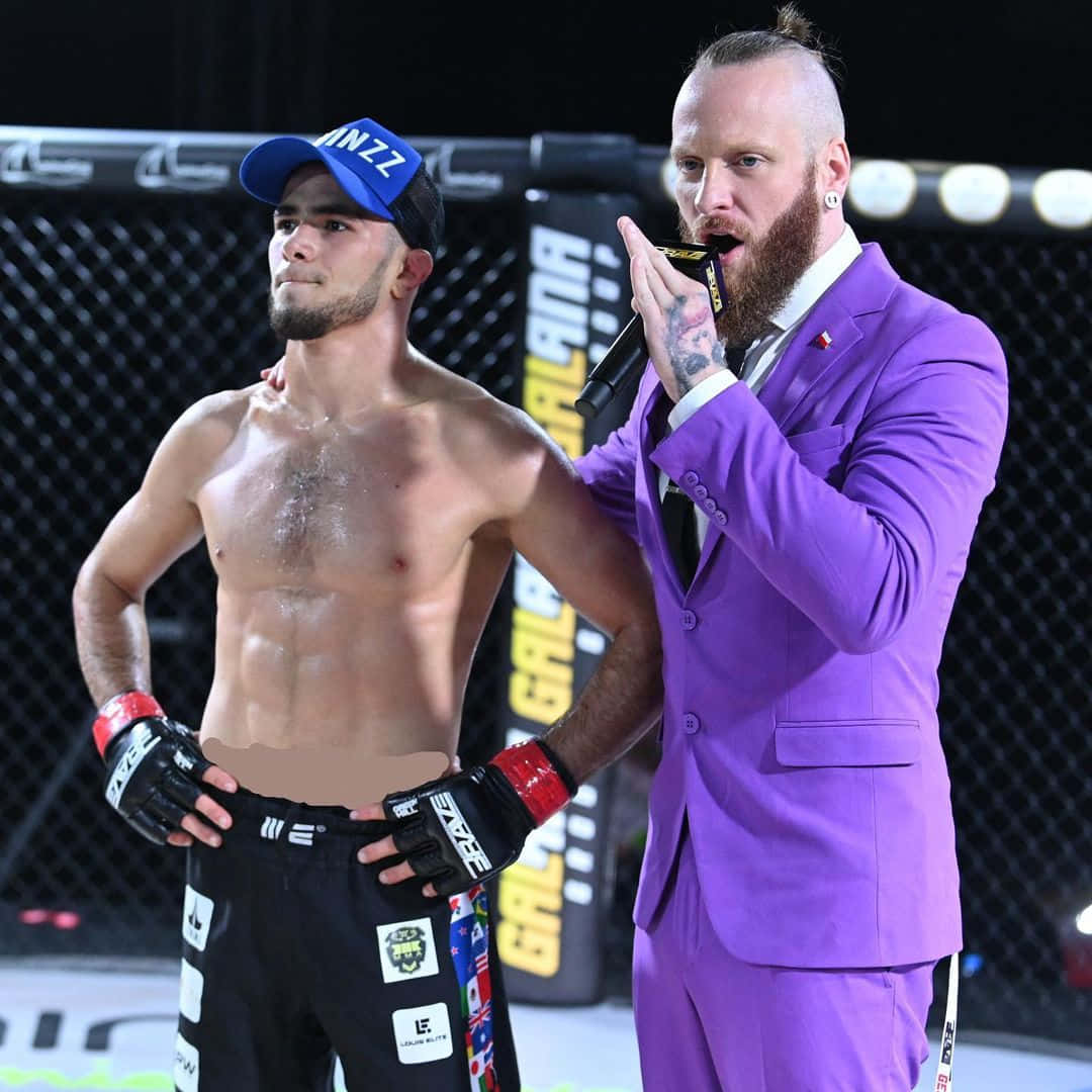 UFC 桌布 壁紙 — 一名肌肉發達的戰士穿著黑色短褲和藍色帽子，站在一位鬍鬚濃密的播音員旁邊，播音員穿著鮮豔的紫色西裝，在籠內拿著麥克風。