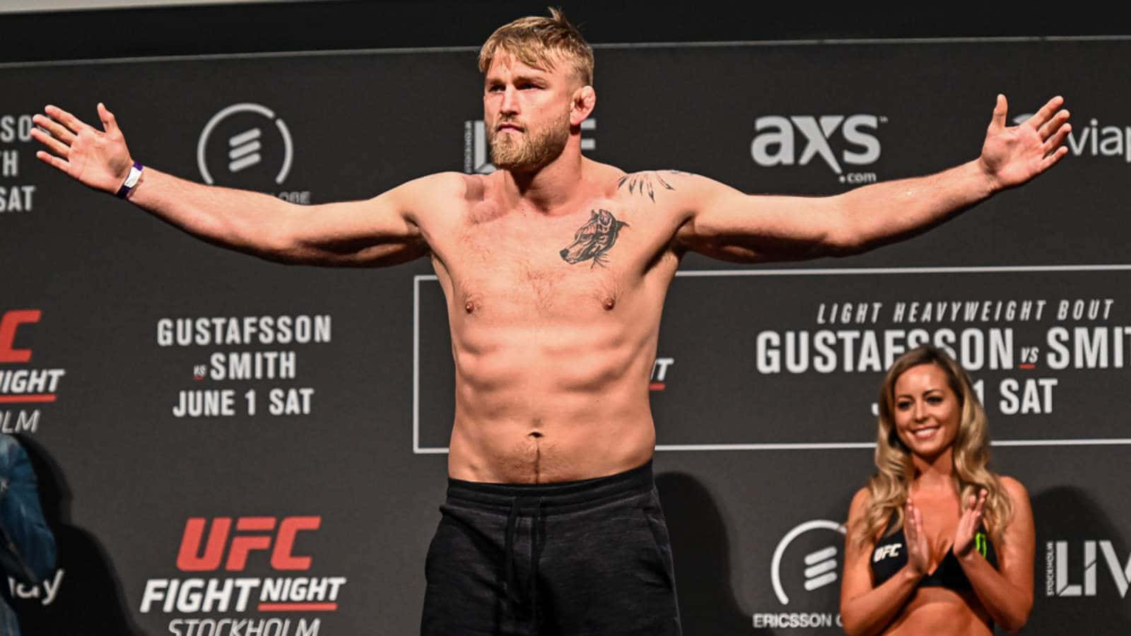UFC 桌布 壁紙 — 一名肌肉發達的戰士赤裸上身，張開雙臂站在UFC稱重台上，旁邊一位微笑的擂台女郎正在鼓掌。