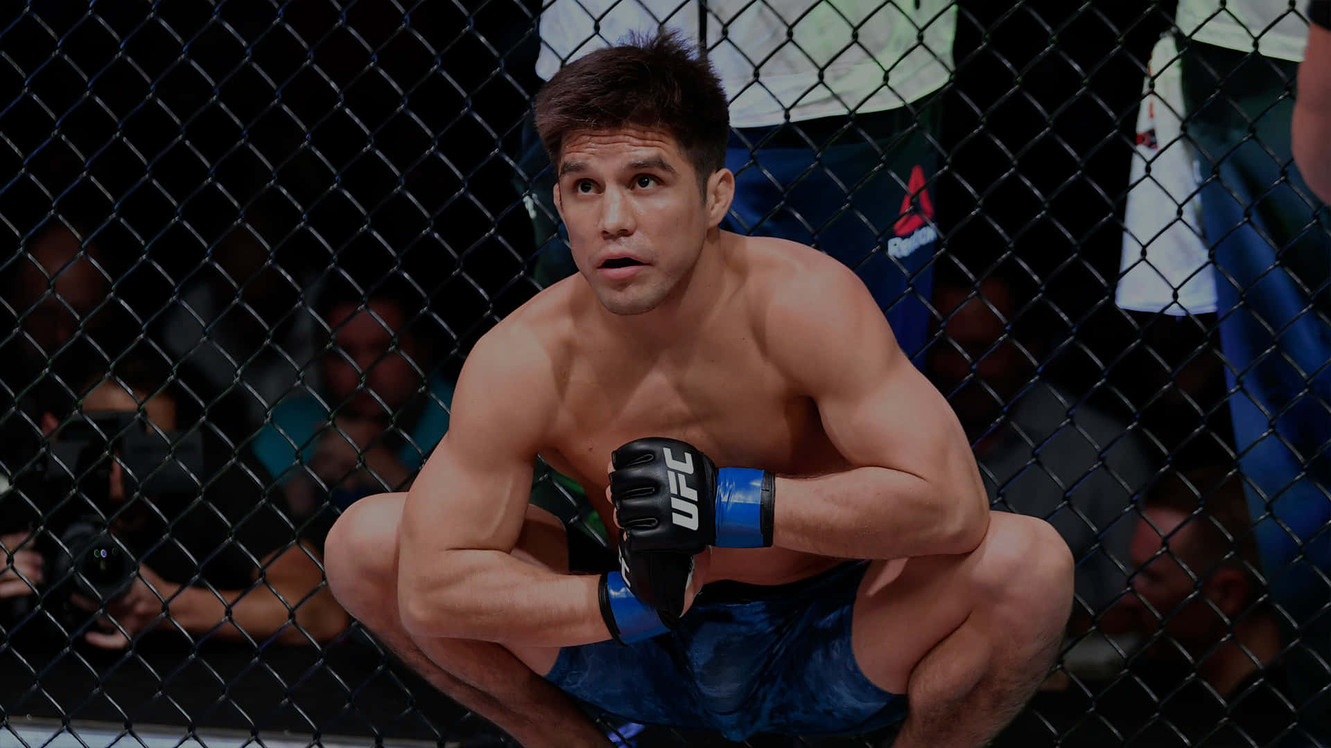 UFC 桌布 壁紙 — 一名肌肉發達的戰士蹲在UFC八角籠內，穿著藍色短褲和黑色手套，神情專注。