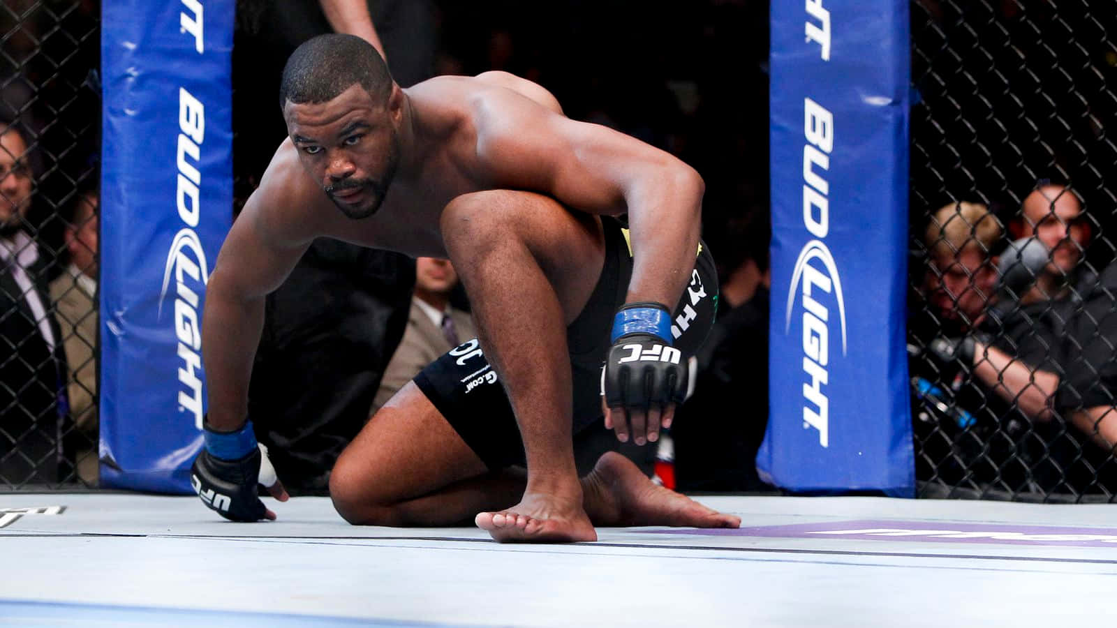 UFC 桌布 壁紙 — 一名肌肉發達的拳手在UFC八角籠中跪下，專注力極強，觀眾在鐵絲網後面觀看。
