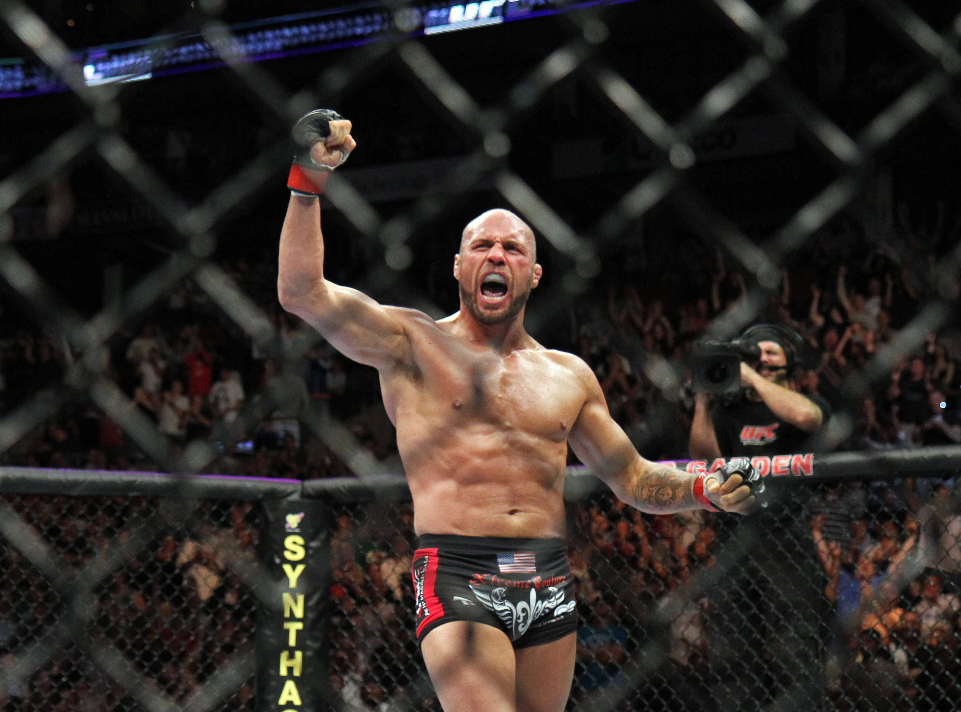 MMA 桌布 壁紙 — 一名肌肉發達的拳手在明亮的場館燈光下，於UFC八角籠內高舉拳頭，發出勝利的怒吼。