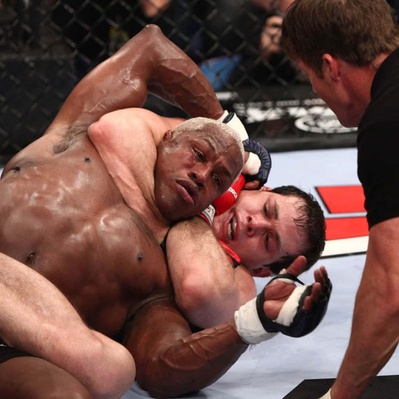 UFC 桌布 壁紙 — 一名肌肉發達的拳手在籠中對對手施展鎖喉，對手痛苦地扭曲表情，裁判在旁緊密觀察。