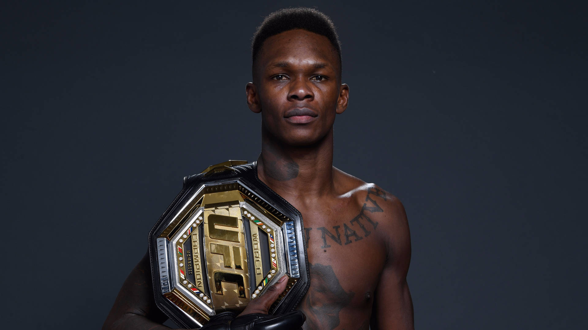 Adesanya 桌布 壁紙 — 一名肌肉發達的拳手在黑暗的攝影棚背景下，以聚焦燈光手持閃亮的UFC冠軍腰帶。