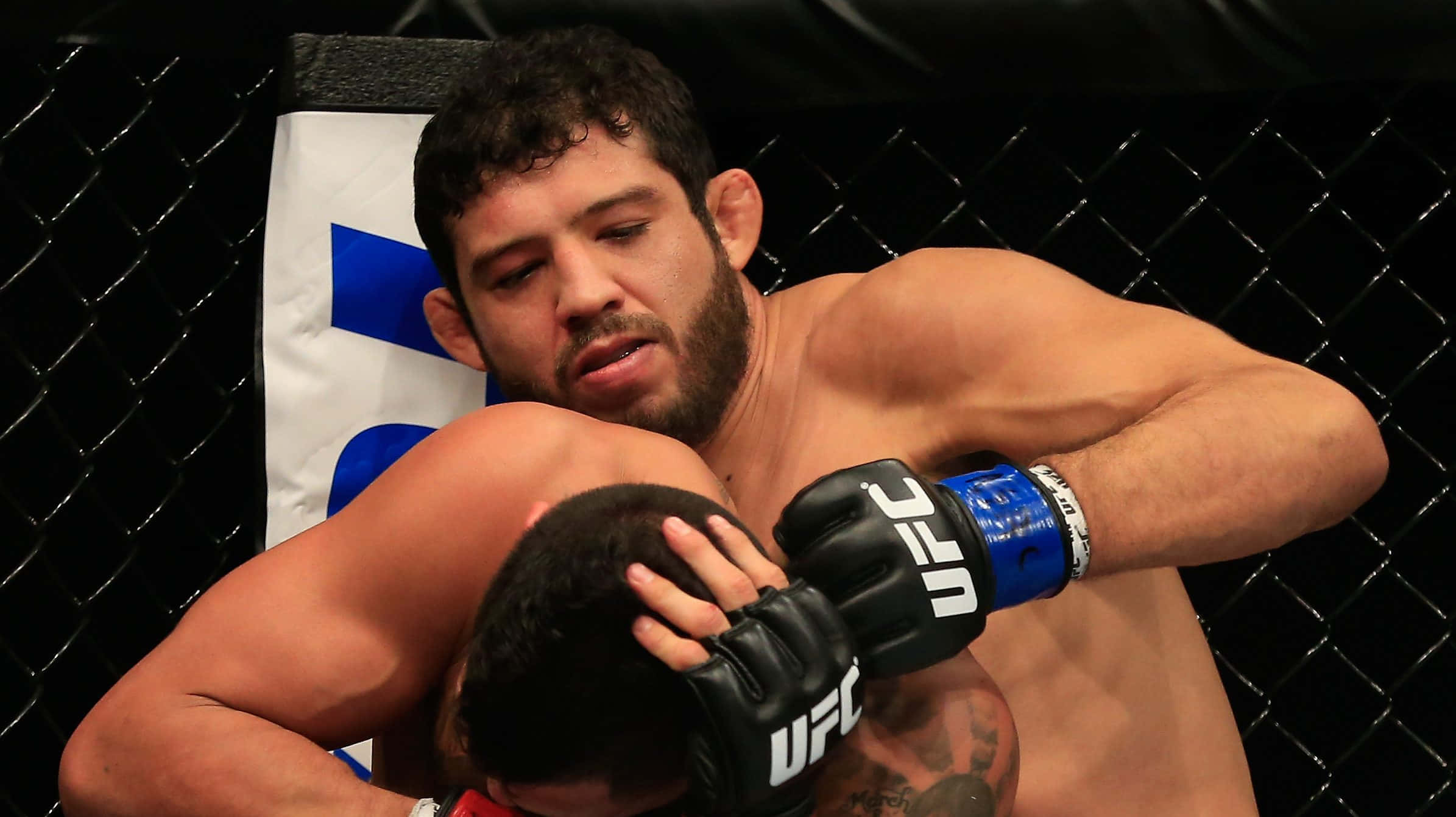 UFC 桌布 壁紙 — 一名肌肉發達的拳手戴著UFC手套，在黑色鐵鏈八角籠內與對手纏鬥。