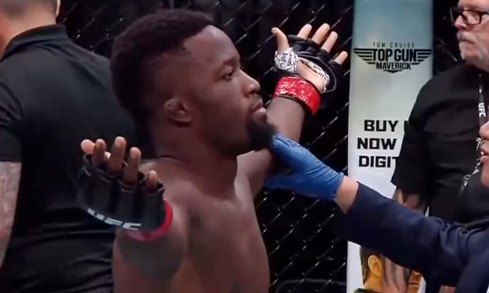 UFC 桌布 壁紙 — 一名肌肉發達的拳手戴著黑色手套在UFC八角籠內高舉雙臂慶祝勝利，裁判戴著藍色手套介入。