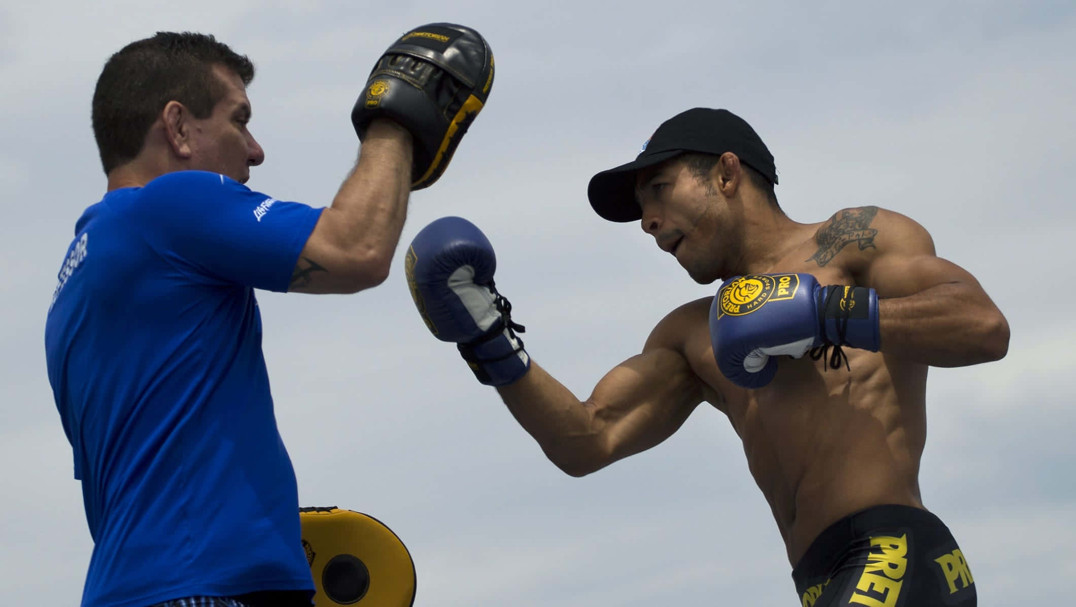 UFC 桌布 壁紙 — 一名肌肉發達的拳擊手戴著藍色手套，在陰天之下向拿著靶子的教練揮拳。