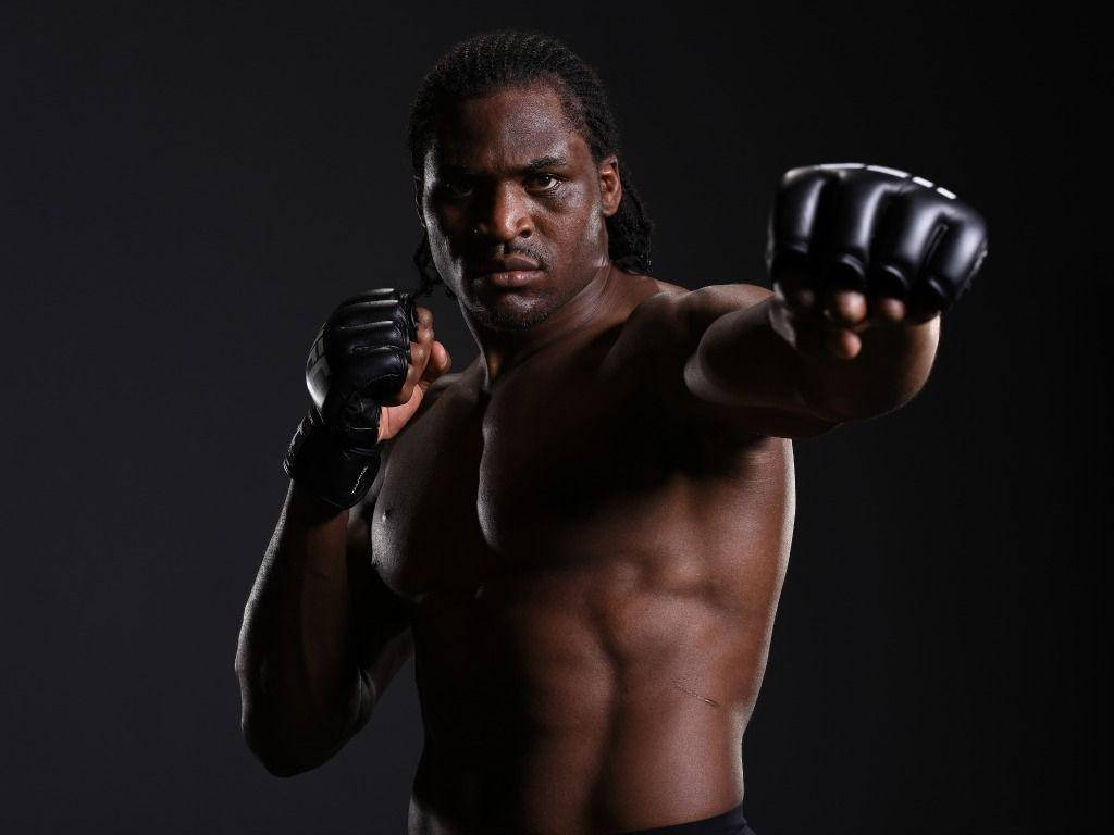 MMA 桌布 壁紙 — 一名肌肉發達的拳擊手戴著黑色手套，在黑暗的攝影棚背景下舉起拳頭，專注地向前凝視。