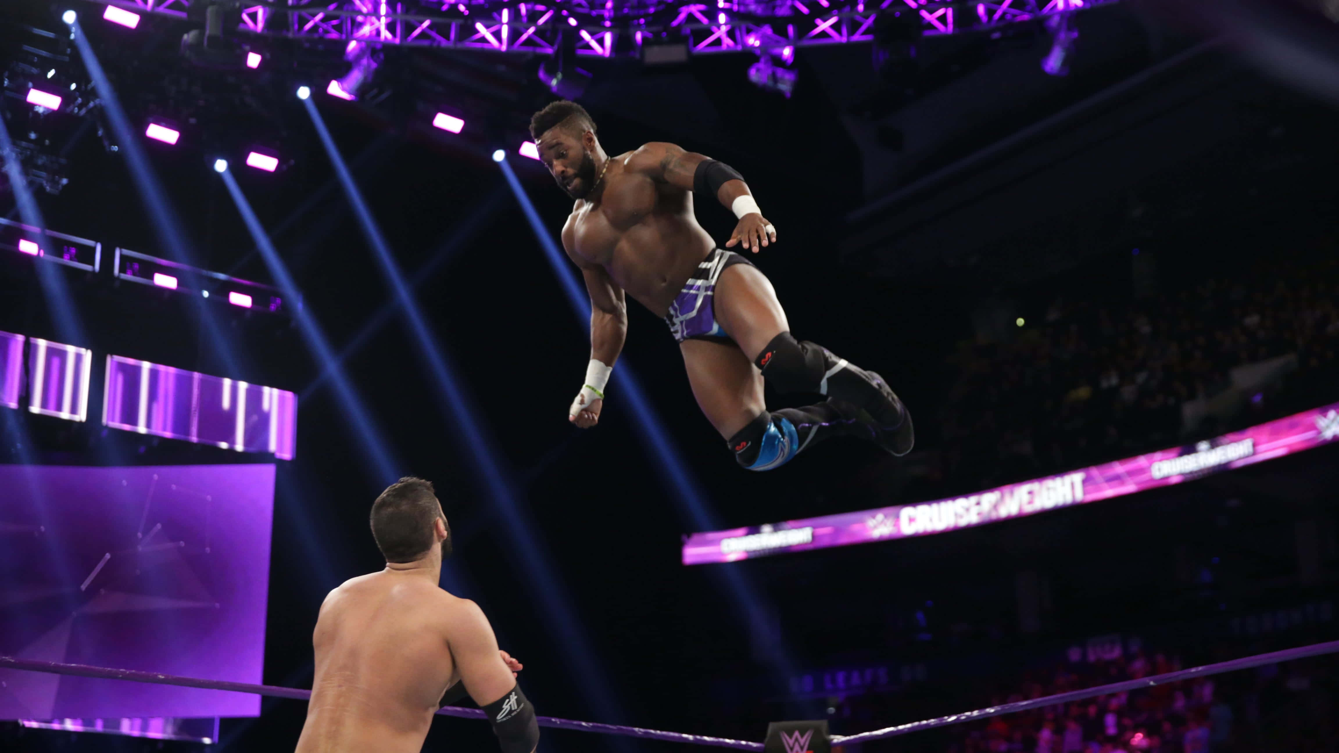 WWE 桌布 壁紙 — 一名肌肉發達的摔角手在紫色舞台燈光下，於明亮照明的擂台上向對手飛身躍起。