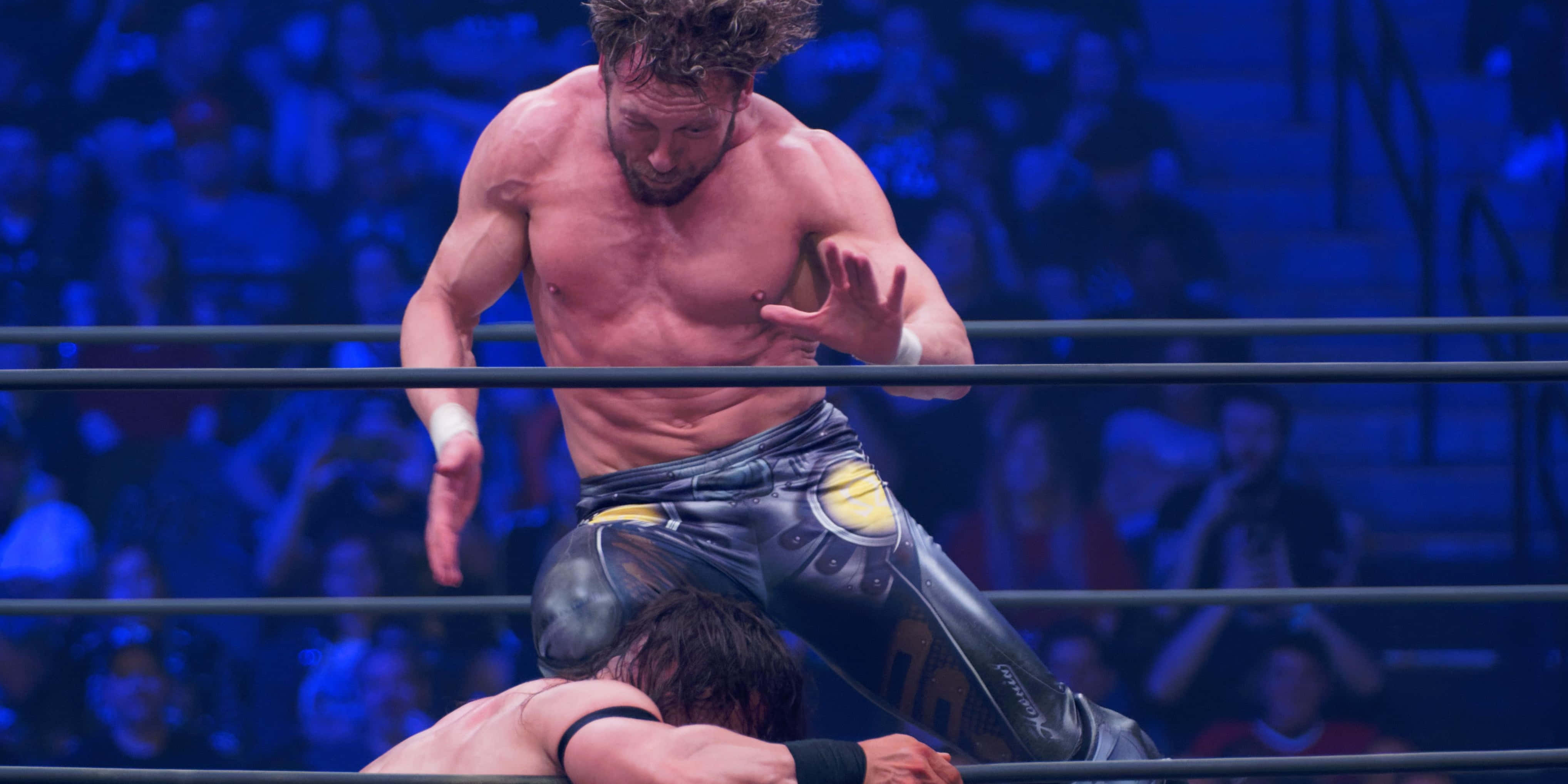 Aew 桌布 壁紙 — 一名肌肉發達的摔角手在藍色場館燈光下跳過擂台上的對手，背景是模糊的人群。