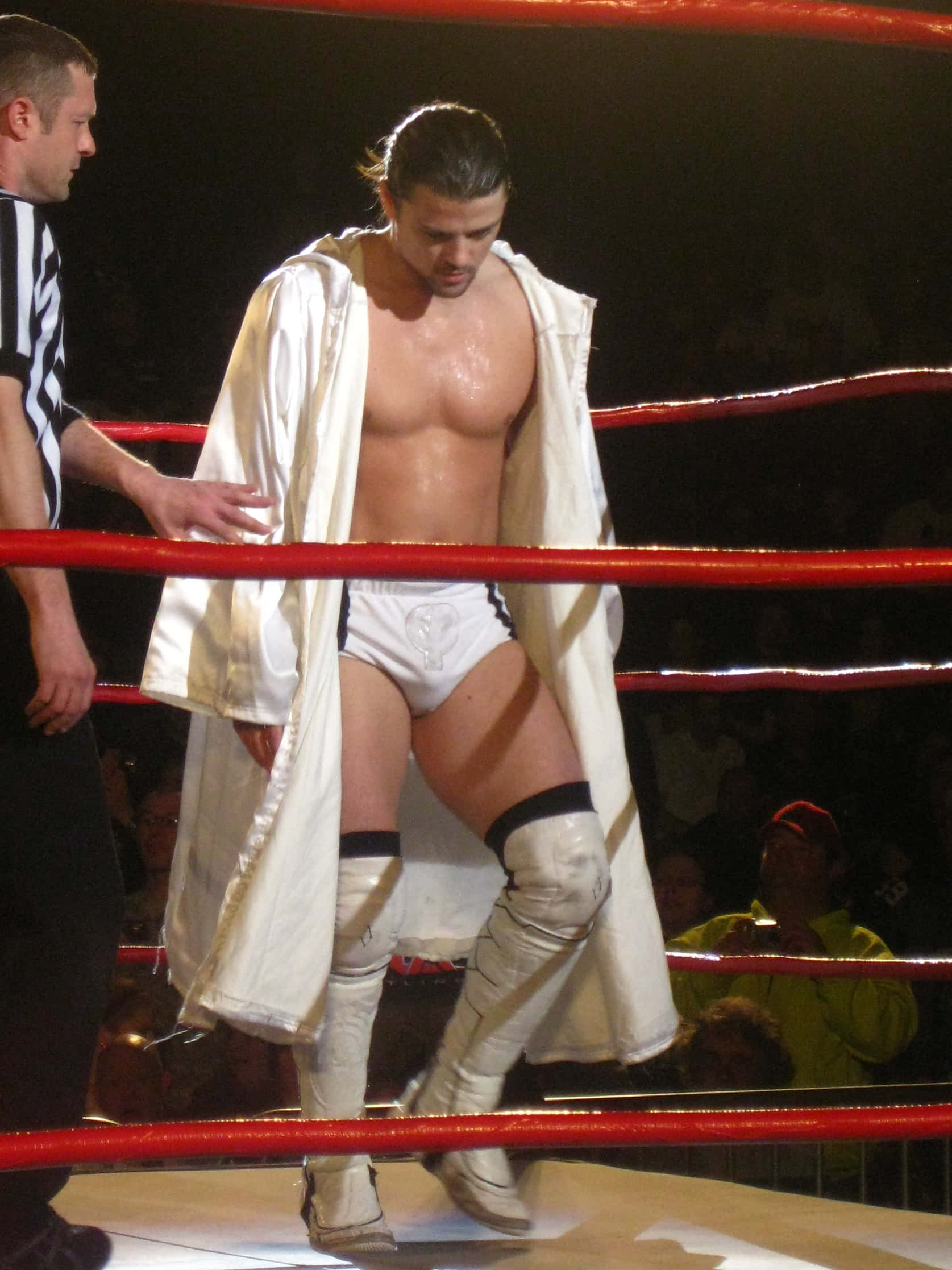 WWE 桌布 壁紙 — 一名肌肉發達的摔角手穿著白色緊身褲和靴子，在明亮的場館燈光下與裁判一起莊嚴地走在擂台上。