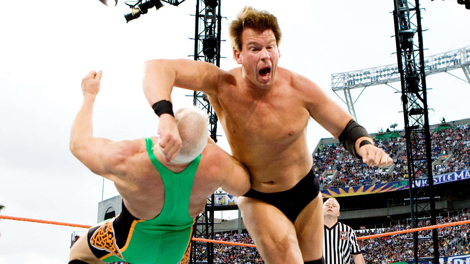 WWE 桌布 壁紙 — 一名肌肉發達的摔角手穿著黑色緊身褲，在明亮的體育館燈光下於WWE比賽中跳起半空，猛擊身穿綠色服裝的對手。