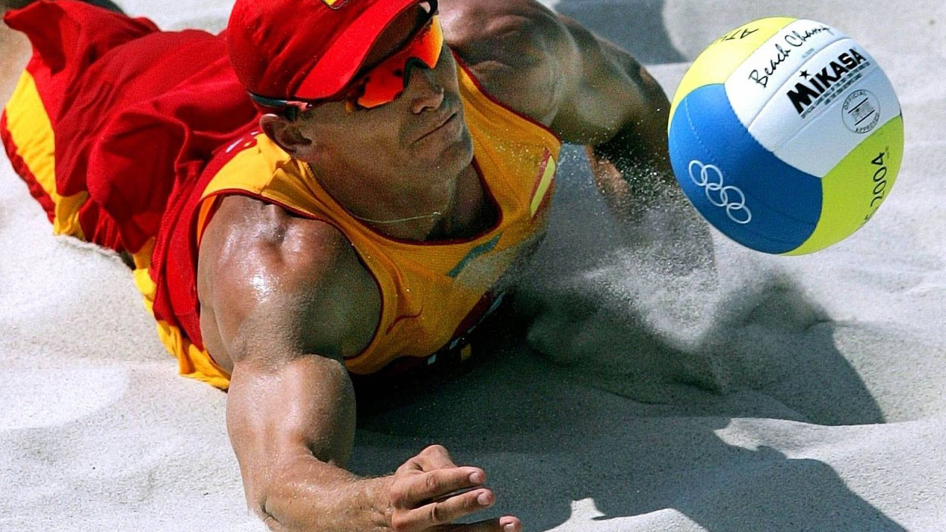 Volleyball 桌布 壁紙 — 一名肌肉發達的沙灘排球運動員，身穿紅黃色裝備，在烈日下潛入沙中扣殺 Mikasa 排球。