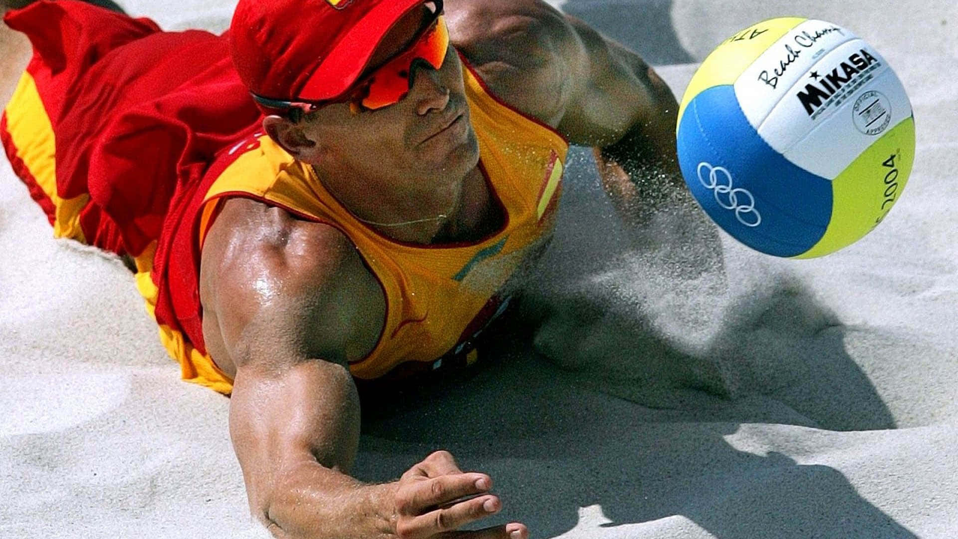 運動 桌布 壁紙 — 一名肌肉發達的沙灘排球運動員身穿紅黃色裝備，在烈日下撲向沙地擊打 Mikasa 排球。