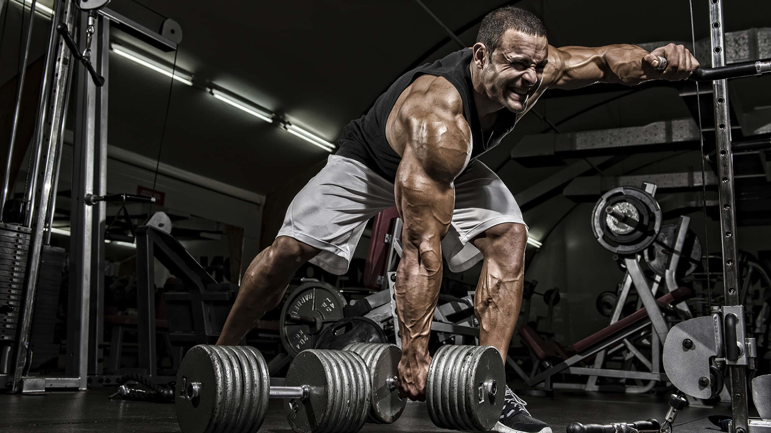 健身房 桌布 壁紙 — 一名肌肉發達的男子在光線昏暗、配備工業設備的健身房中，用力舉起沉重的槓鈴。