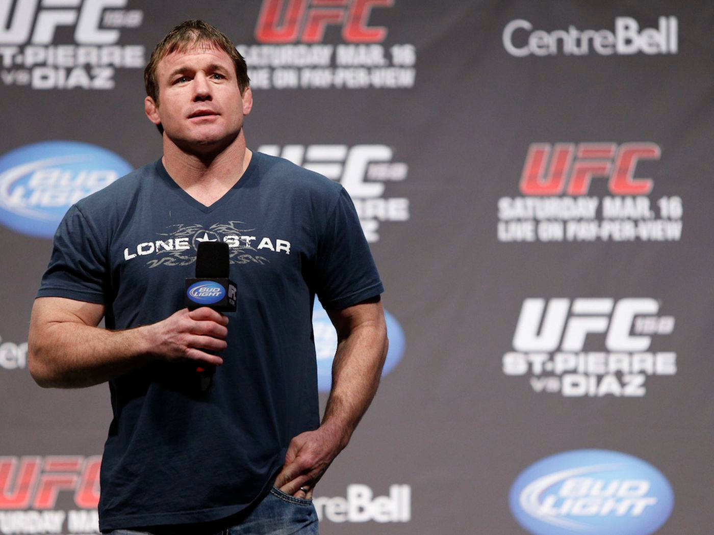 UFC 桌布 壁紙 — 一名肌肉發達的男子身穿藍色Lone Star T恤，在帶有品牌背景的UFC新聞發布會上手持麥克風。