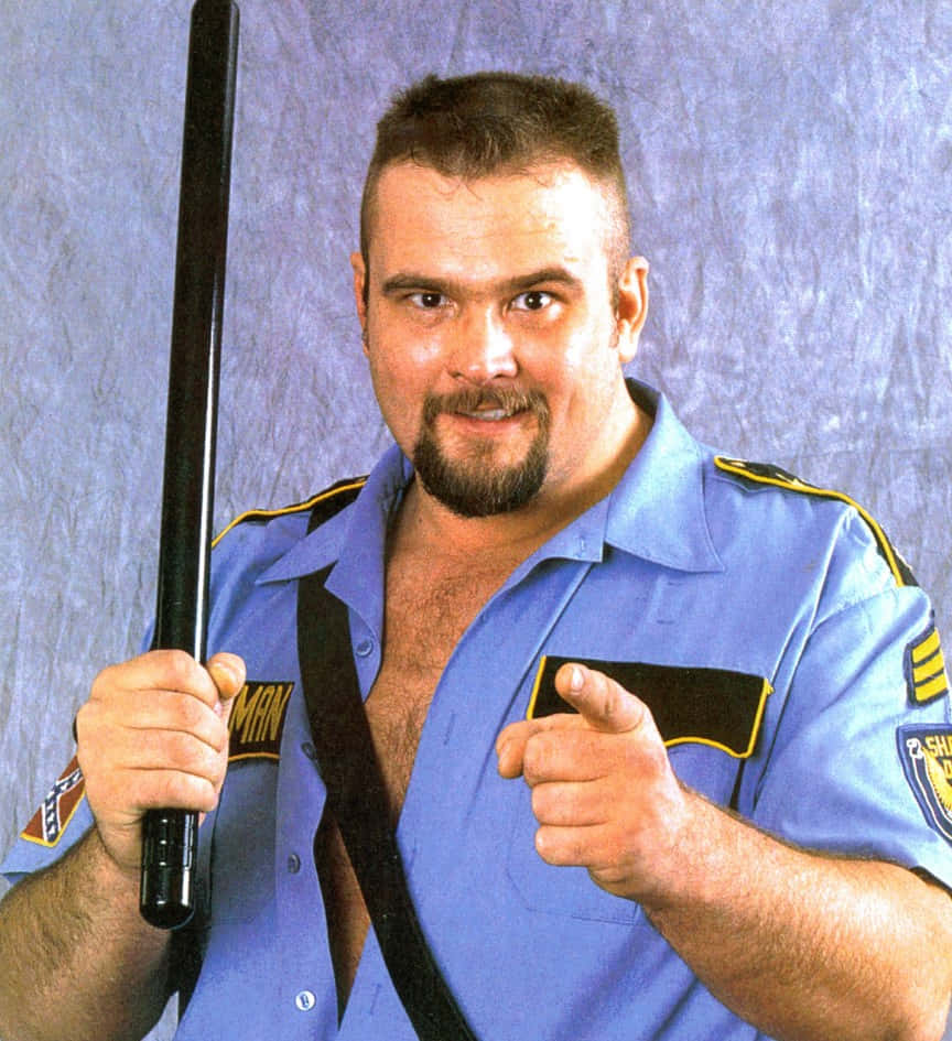 Big Boss Man 桌布 壁紙 — 一名肌肉發達的男子身穿藍色警察制服，手持警棍，自信地微笑並直指鏡頭。