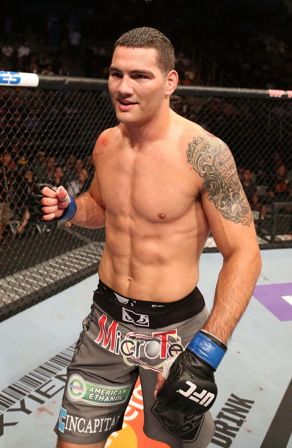 MMA 桌布 壁紙 — 一名肌肉發達的男性拳手，手臂有刺青，自信地站在UFC八角籠內，戴著手套和穿著品牌短褲。
