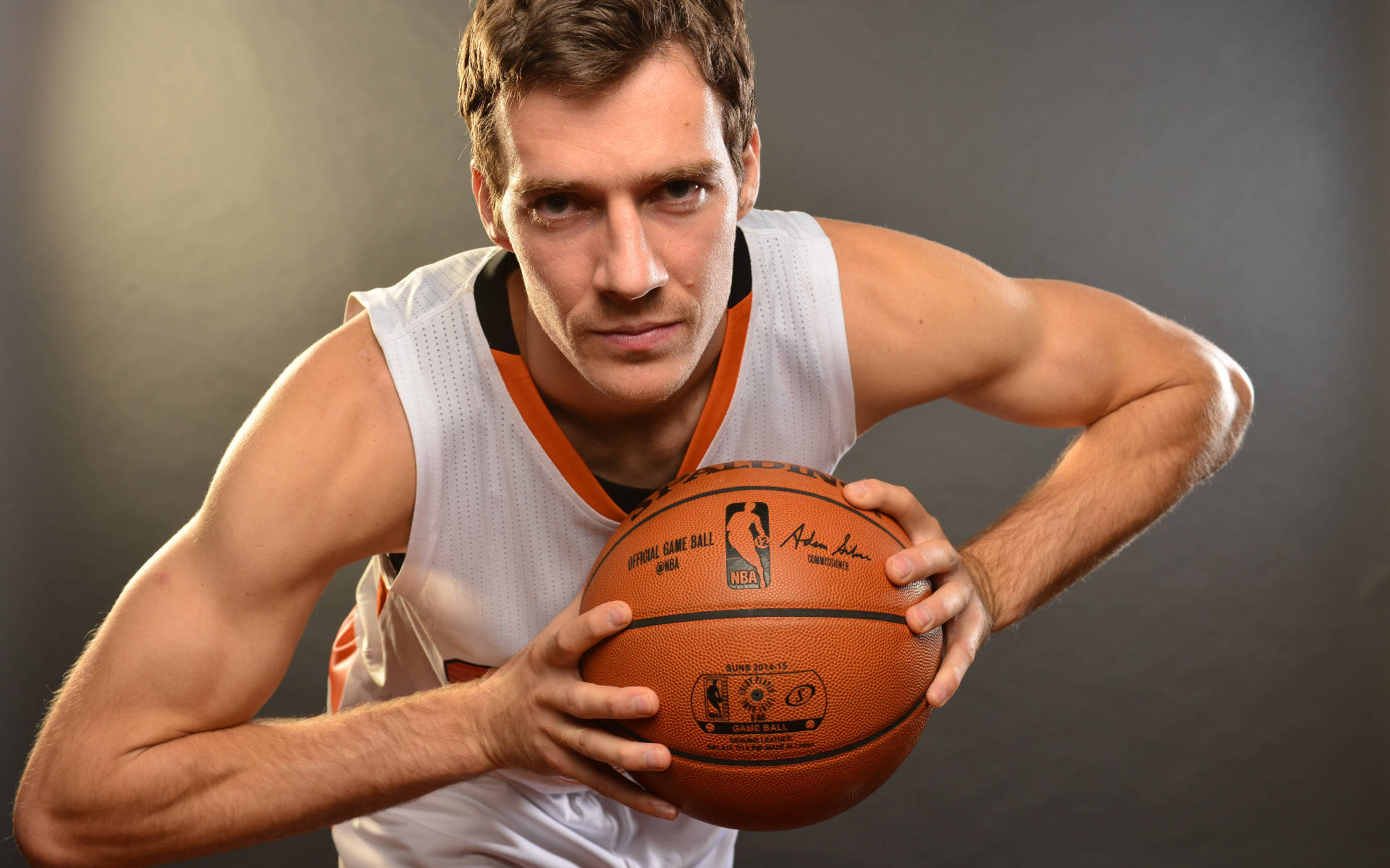Suns 桌布 壁紙 — 一名肌肉發達的籃球運動員身穿白色和橙色球衣，在深色工作室背景下，專注地緊握籃球。