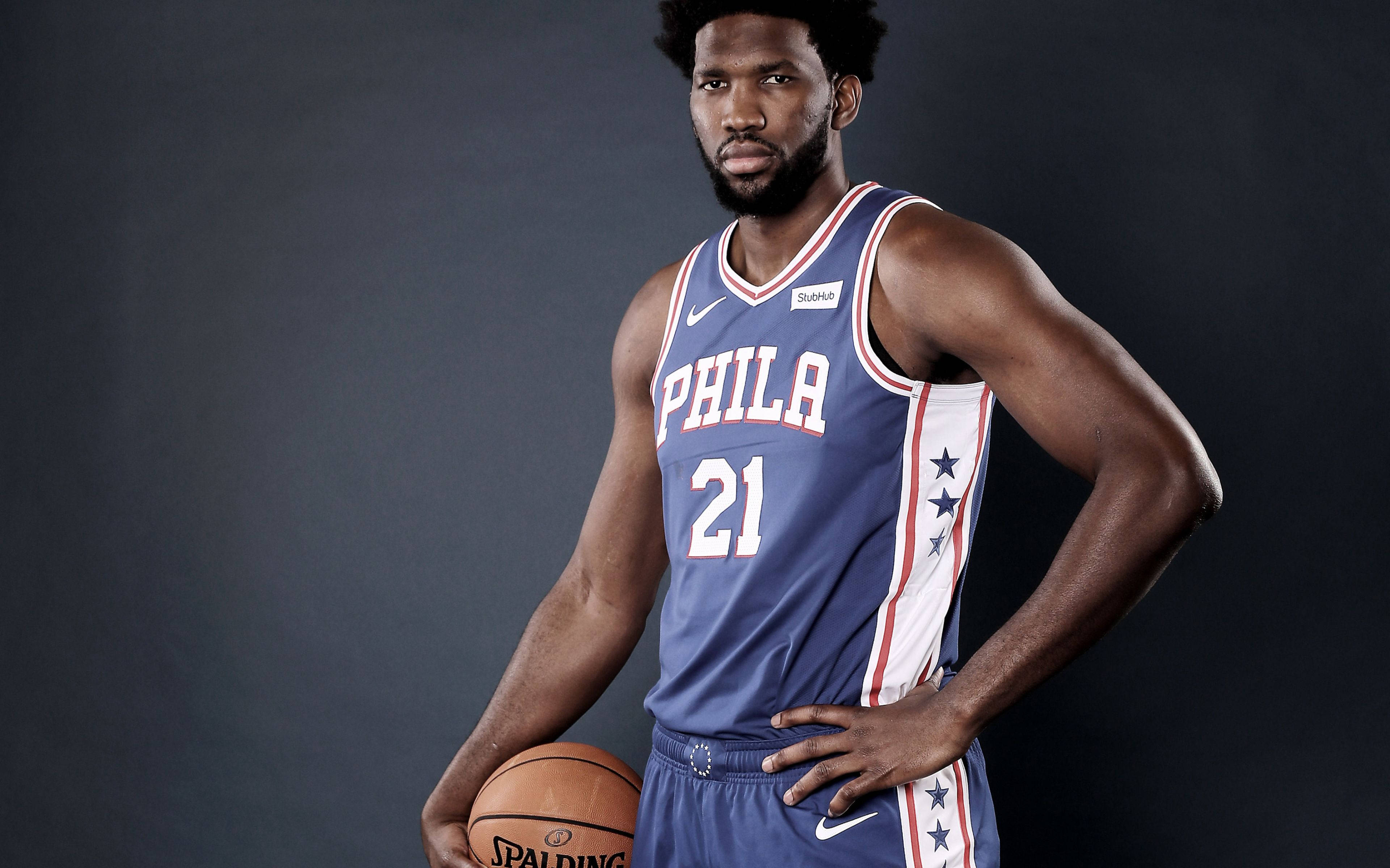 Embiid 桌布 壁紙 — 一名肌肉發達的籃球運動員身穿藍白紅費城76人球衣，在黑暗的攝影棚背景下拿著一個Spalding籃球。