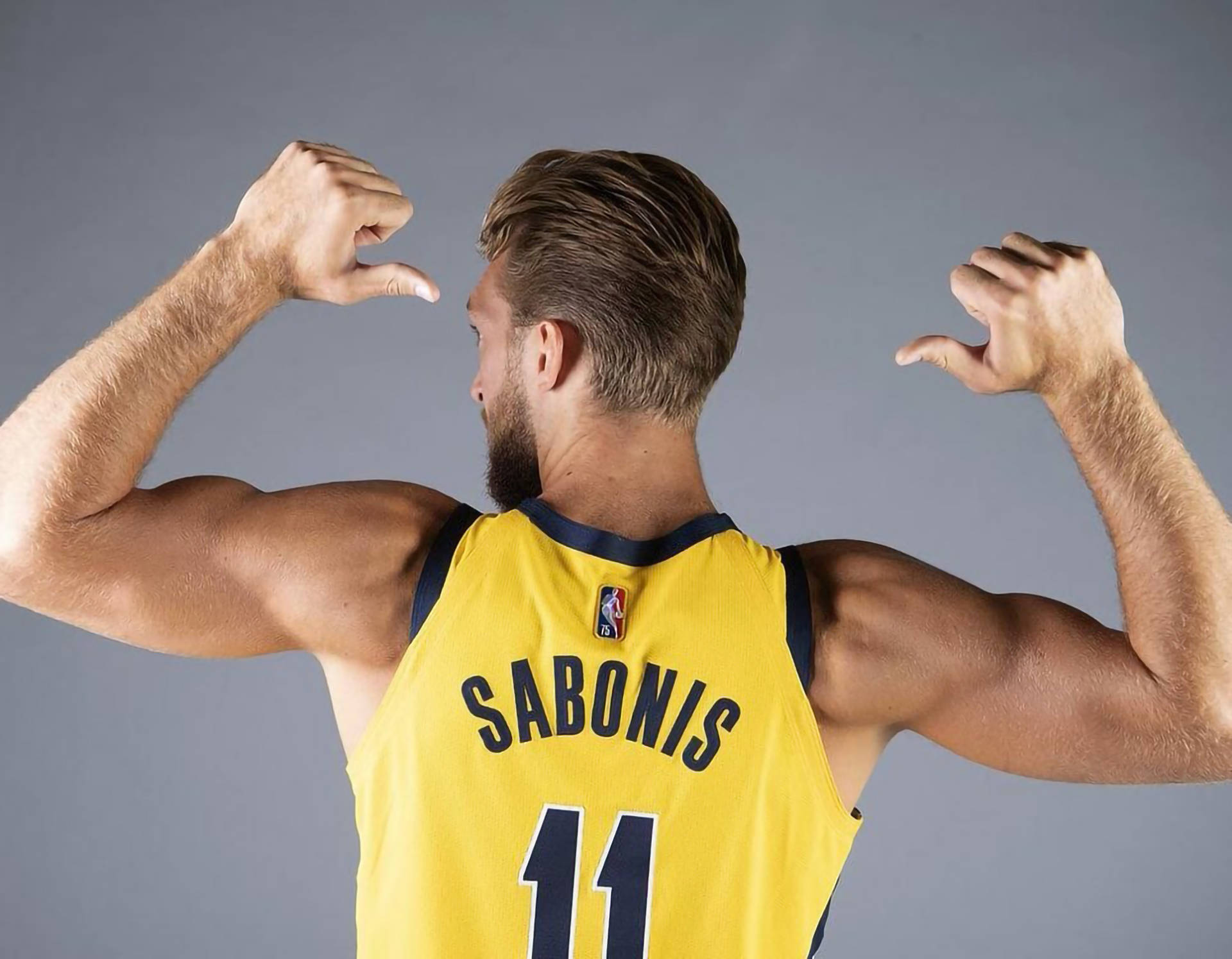 domantas sabonis 桌布 壁紙 — 一名肌肉發達的籃球運動員身穿黃色球衣，背對鏡頭，在灰色工作室背景前彎曲手臂。