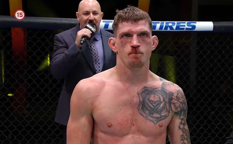 UFC 桌布 壁紙 — 一名胸口有刺青、血跡斑斑的MMA拳手站在八角籠內，身後有位評論員正對著麥克風說話。
