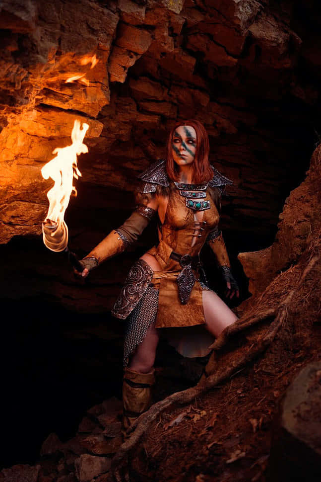 Aela The Huntress 桌布 壁紙 — 一名臉上塗有藍色顏料的戰士在黑暗的岩石洞穴中手持燃燒的火把，照亮了崎嶇的石牆。