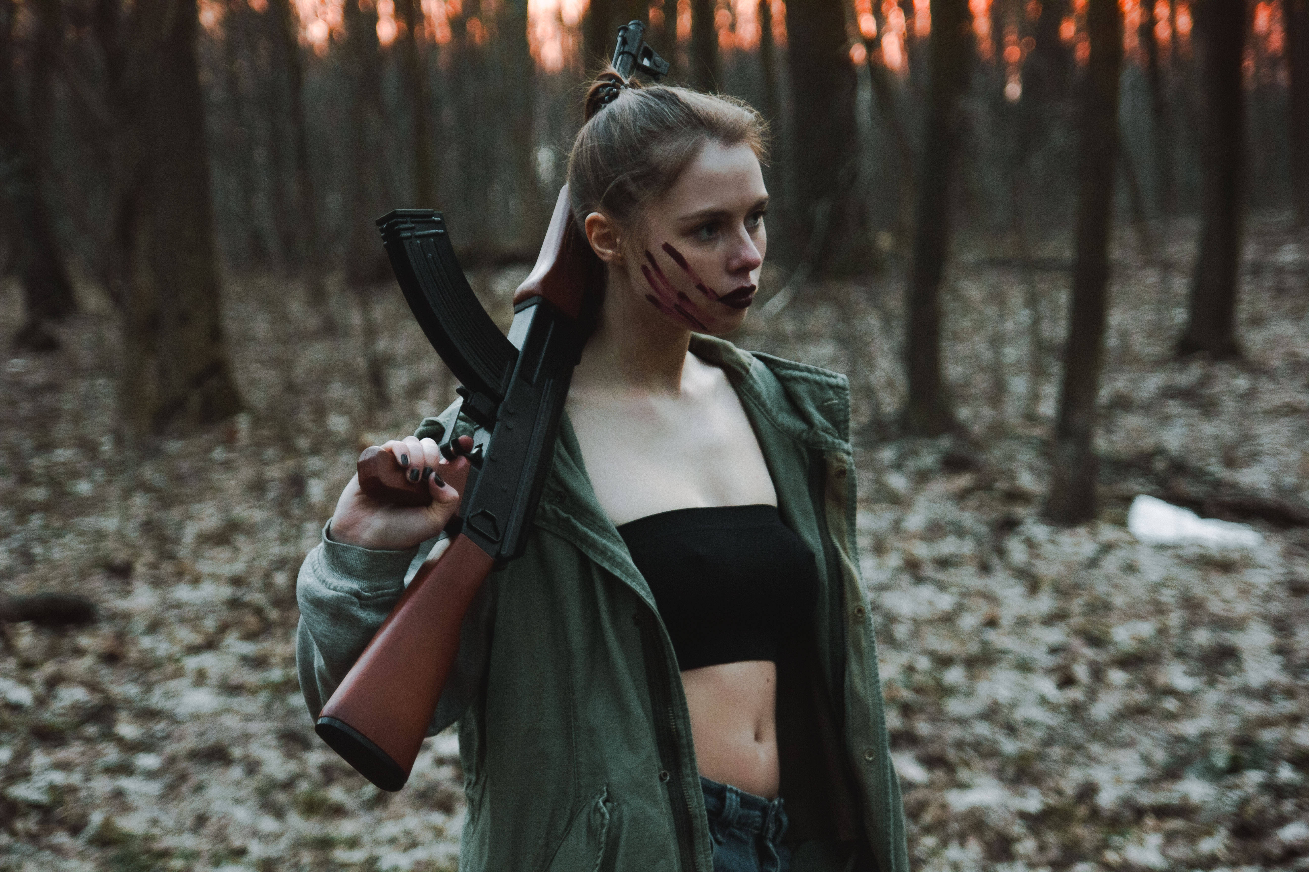Warrior 桌布 壁紙 — 一名臉上帶有血痕的女子在黃昏時分的昏暗森林中手持突擊步槍。