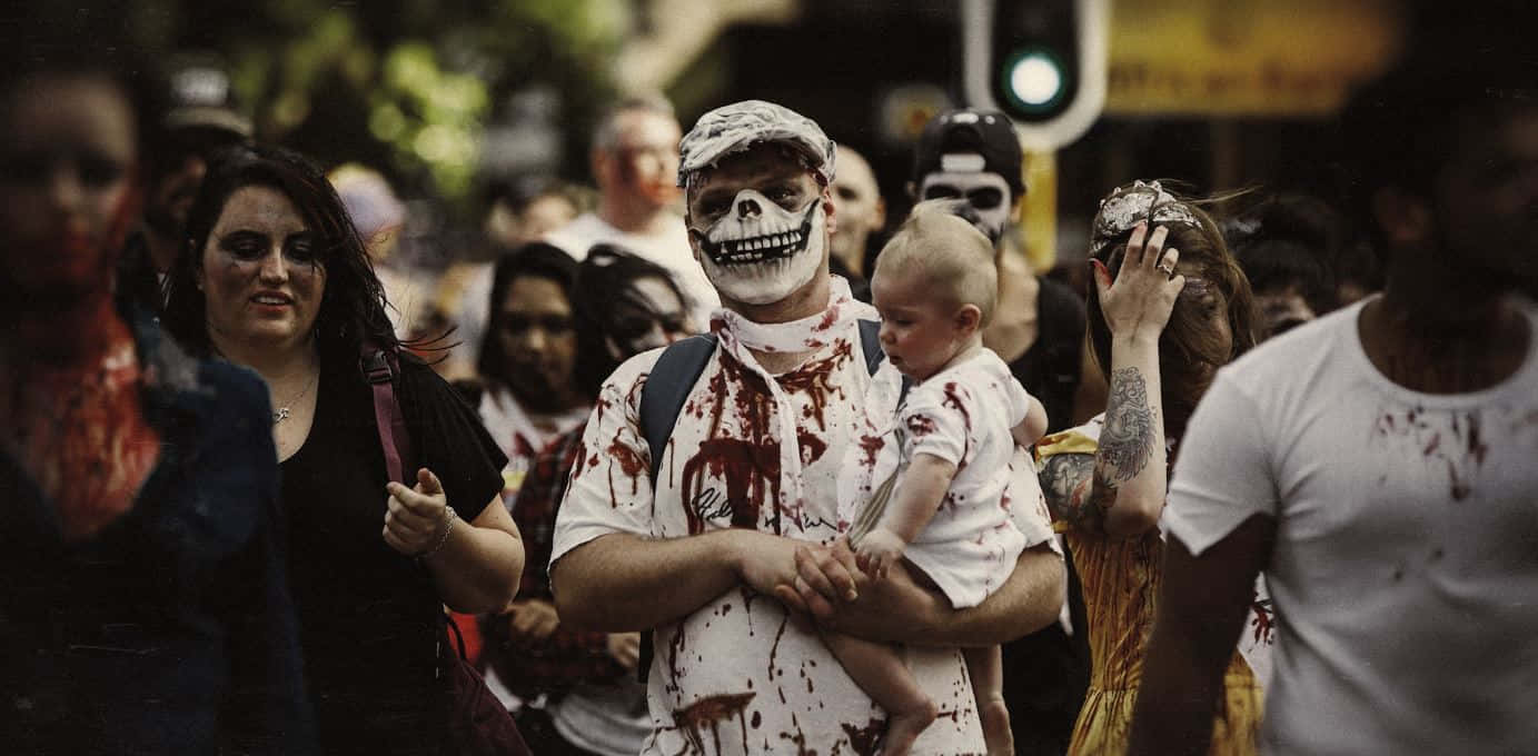 Halloween Parades 桌布 壁紙 — 一名臉上畫著骷髏妝的男子，在擠滿人群的殭屍主題街頭遊行中，抱著一個沾滿假血的嬰兒。
