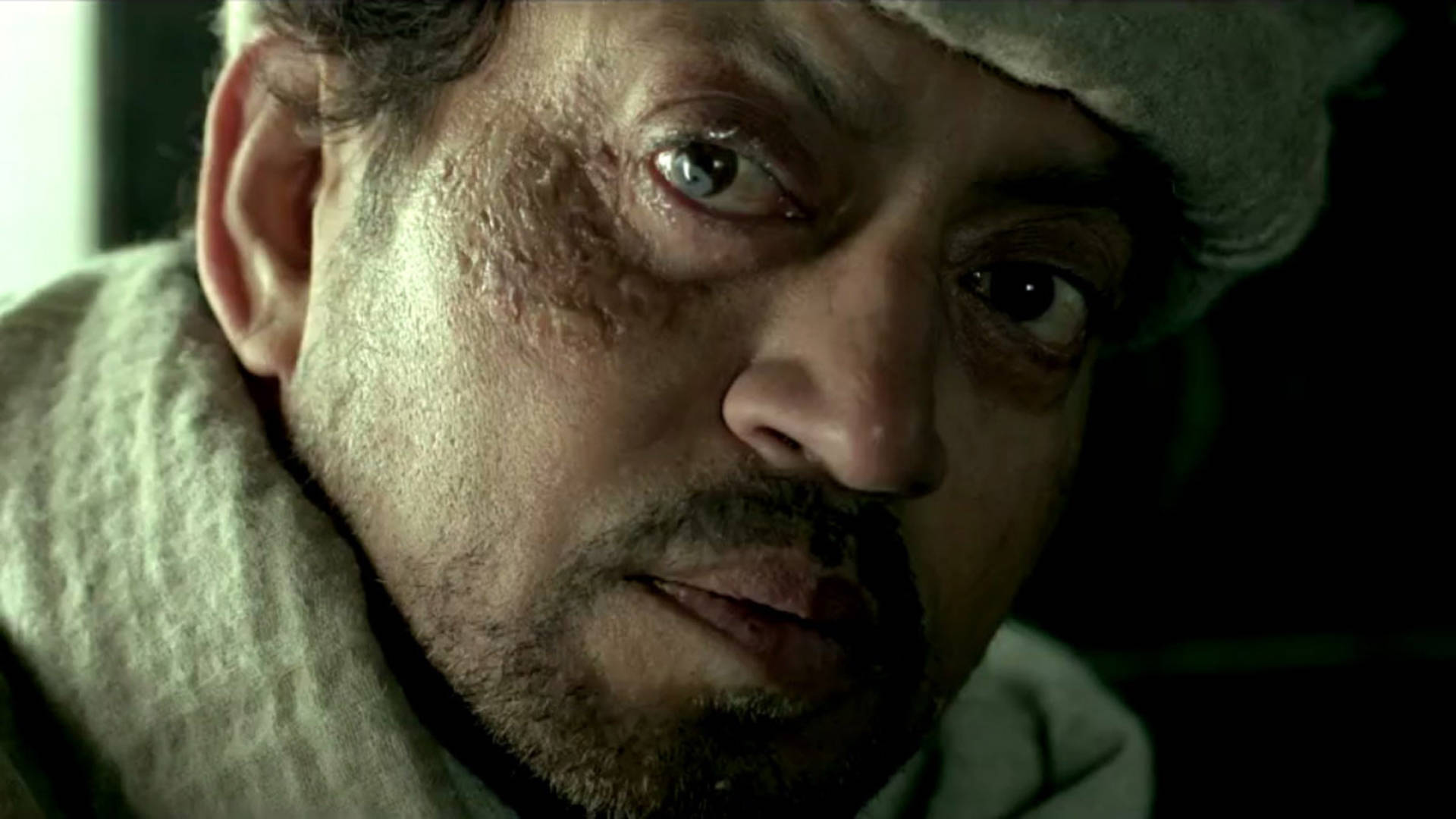 Irrfan Khan 桌布 壁紙 — 一名臉頰有疤痕、眼神銳利的男子，在昏暗燈光下戴著羊毛帽和圍巾的特寫。