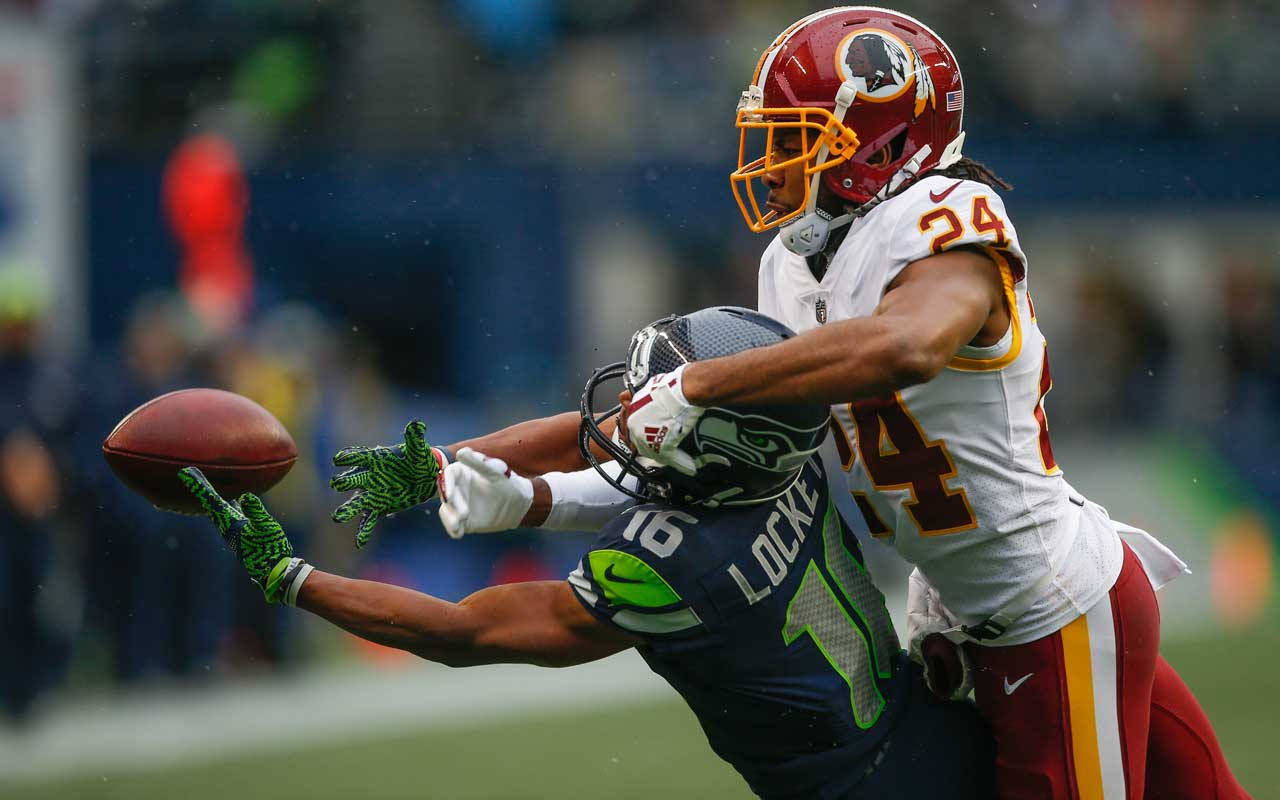 Seattle Seahawks 桌布 壁紙 — 一名華盛頓美式足球隊球員，戴紅色頭盔、穿白色球衣，跳起接球，同時被西雅圖海鷹隊藍色球衣的防守球員擒抱。
