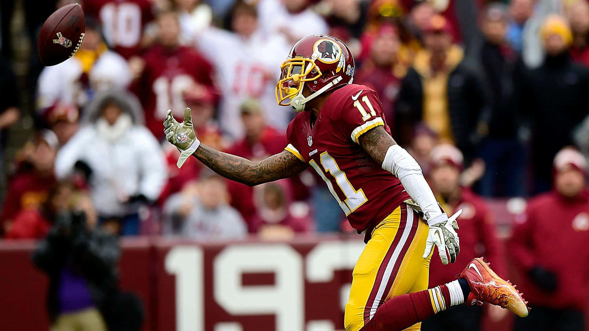 Sean Taylor 桌布 壁紙 — 一名華盛頓美式足球隊的球員身穿棗紅色和金色球衣，在日間比賽中跳起接住美式足球，背景有模糊的球迷。