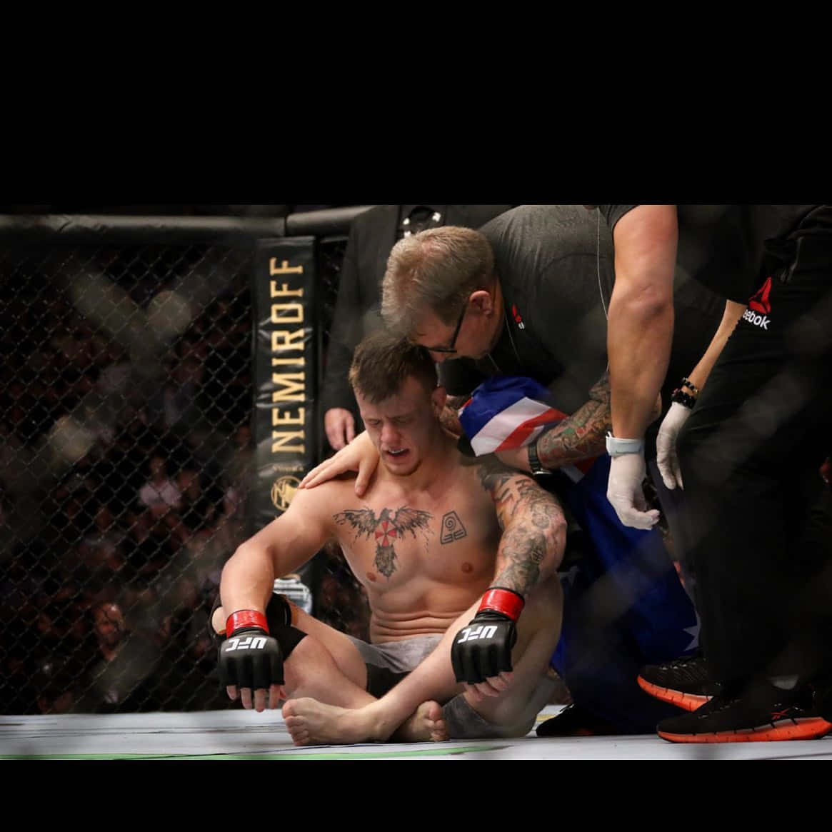 UFC 桌布 壁紙 — 一名落敗的MMA拳手坐在八角籠地上，醫護人員在明亮的場館燈光下為他治療。