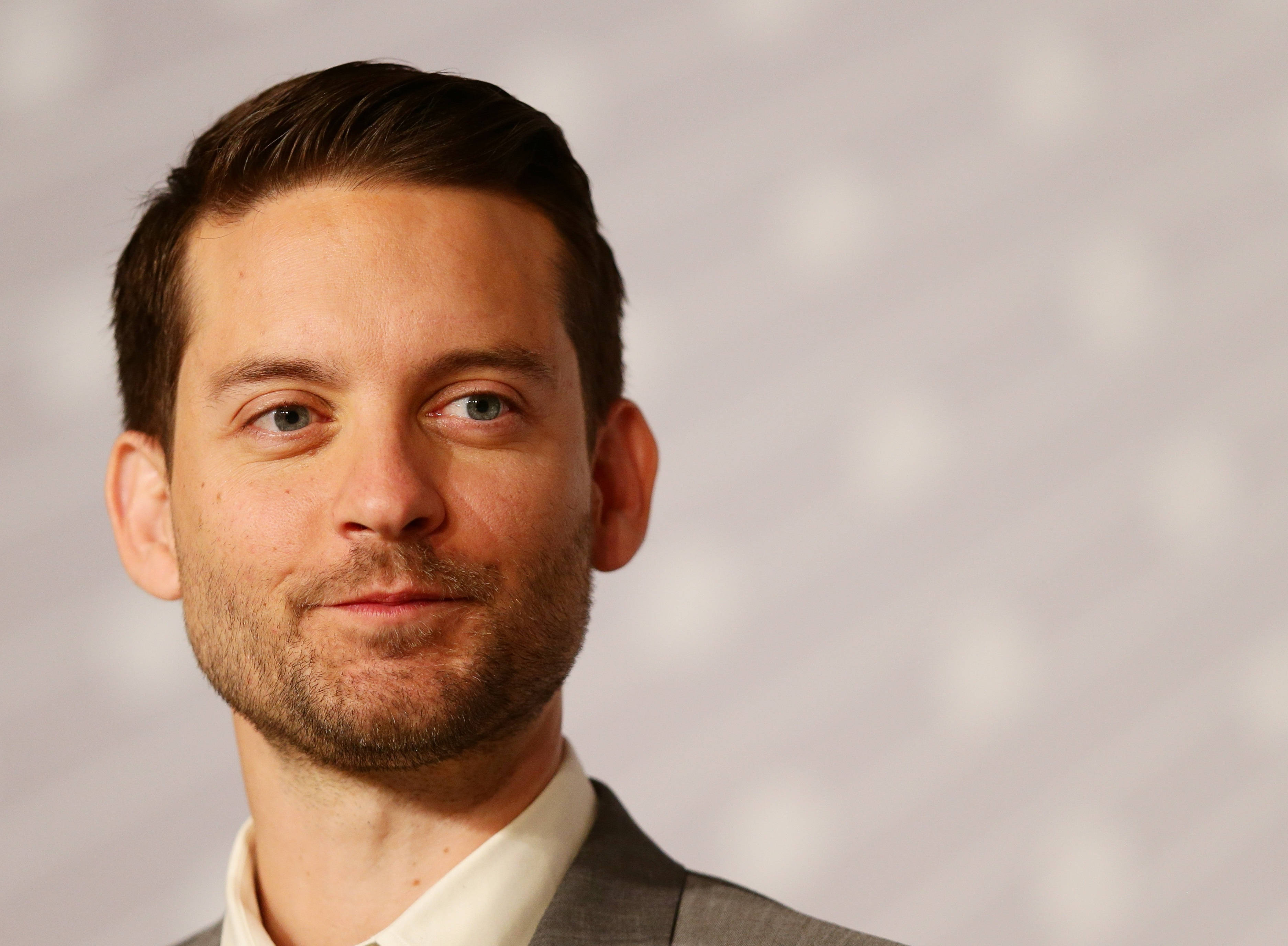 Tobey Maguire 桌布 壁紙 — 一名藍眼睛、短棕髮男士的 close-up 肖像，身穿灰色西裝，背景為柔軟的白色紋理背景。