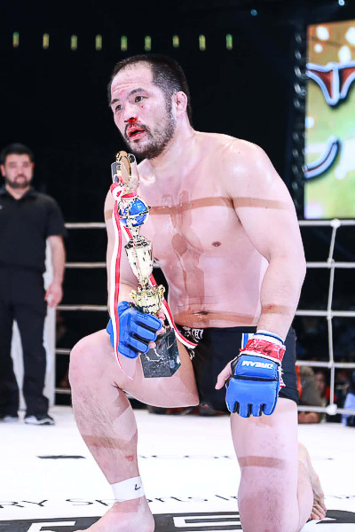 MMA 桌布 壁紙 — 一名血跡斑斑的MMA拳手在八角籠中跪地，手持冠軍獎盃，戴著藍色手套，軀幹上纏著膠帶。