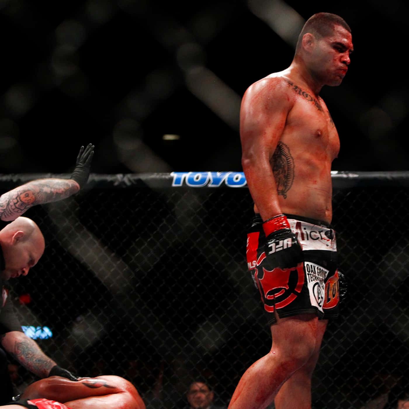 UFC 桌布 壁紙 — 一名血跡斑斑的MMA拳手在八角籠中勝利站立，而他的對手在明亮的場館燈光下跪地敗北。