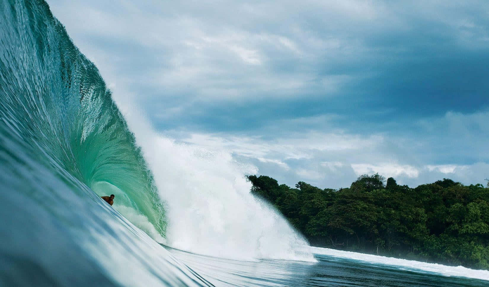 surfing 桌布 壁紙 — 一名衝浪者在多雲的天空下，騎乘一道巨大的碧綠色海浪，海浪翻捲在鬱鬱蔥蔥的海岸線上。