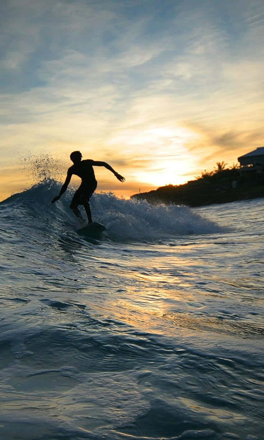 Surfing iPhone 桌布 壁紙 — 一名衝浪者在日落時分乘風破浪，金色光線在水面上反射，剪影映襯著色彩繽紛的天空。
