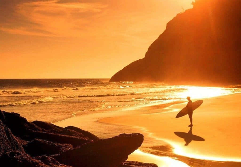 surfing 桌布 壁紙 — 一名衝浪者在日落時分沿著金色沙灘漫步，海浪拍打，懸崖呈現剪影。