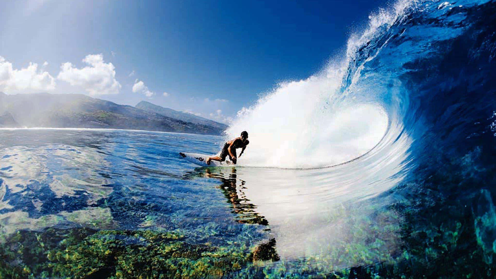 surfing 桌布 壁紙 — 一名衝浪者在明亮的藍天下騎乘巨大的藍綠色海浪，下方清晰可見海底礁石。