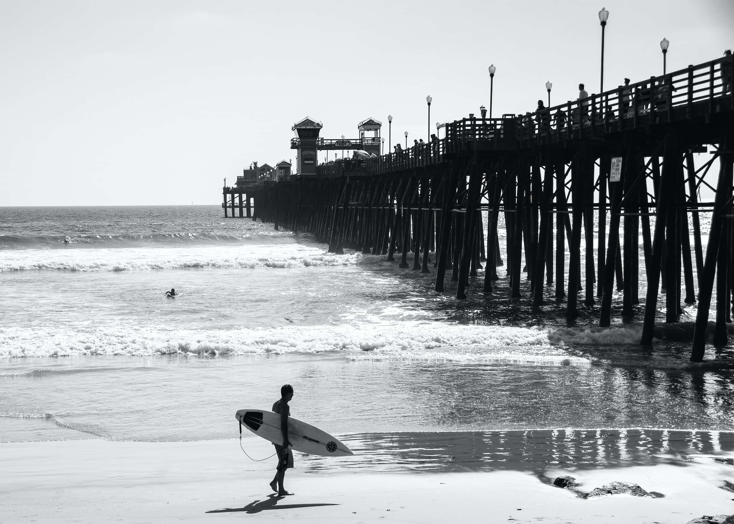 Black And White Beach 桌布 壁紙 — 一名衝浪者在陽光明媚的海灘上行走，帶著一塊衝浪板，海浪在長木碼頭附近拍打。